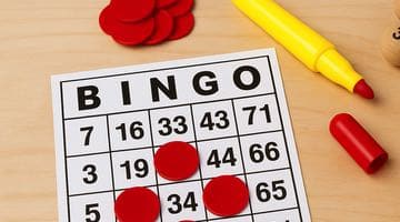 En bingobricka med röda markörer placerade på siffror. Bredvid spelbrickan ligger nummermarkörer och en gul penna.