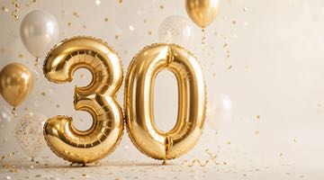Guldiga ballonger formade som siffran 30, omgivna av konfetti och festballonger mot en ljus bakgrund.